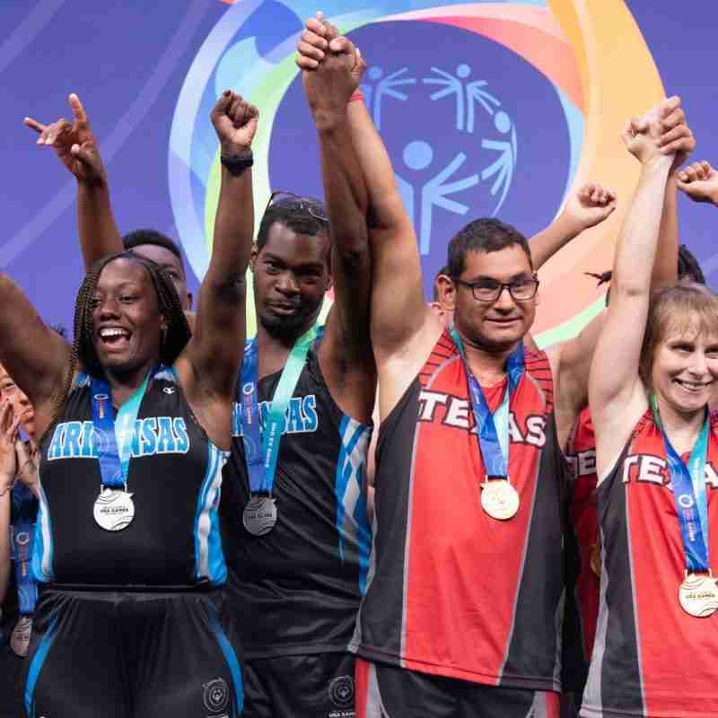 Four individuals wearing gold medals with blue ribbons stand hand in hand with their arms raised in front of a purple background with the Special Olympics USA Games logo.